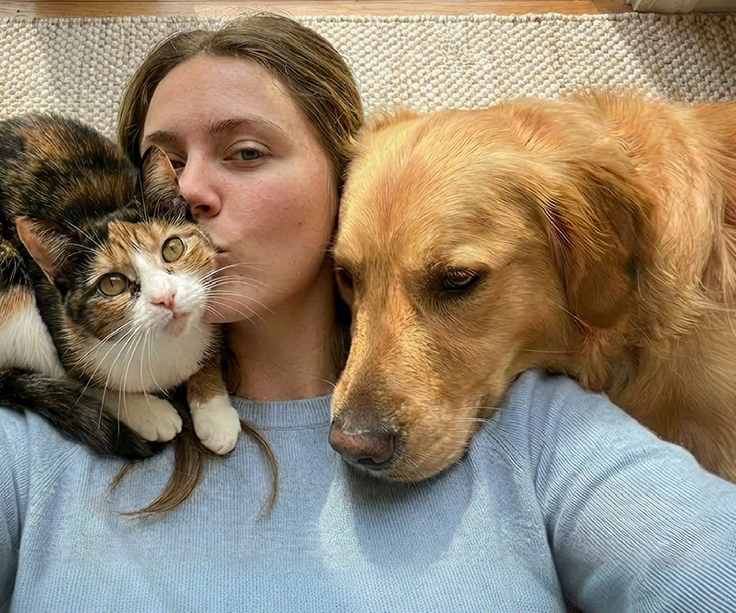 Frau liegt auf dem Boden und kuschelt sowohl mit ihrer Katze als auch ihrem Hund.